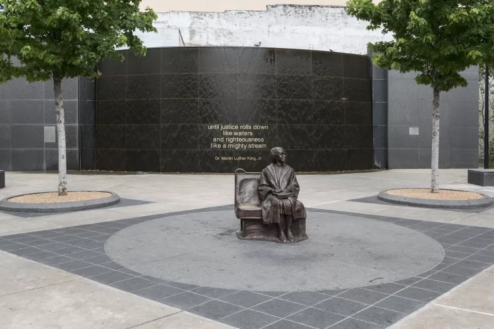 Bronze statue of Rosa Parks sitting on bus bench in center of plaza by Dallas transit station.